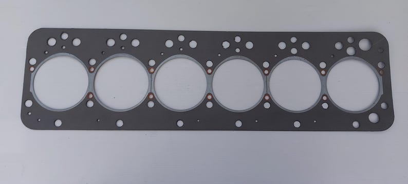 Puede incluir: Una junta de culata rectangular gris con seis grandes aberturas circulares y numerosos agujeros m&aacute;s peque&ntilde;os. La junta tiene un borde gris claro alrededor de las grandes aberturas y detalles en color cobre.