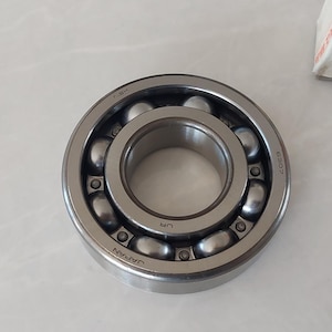 May include: A close-up of a silver ball bearing with multiple small ball bearings inside. The bearing is on a white surface. A white and red label is partially visible in the background.