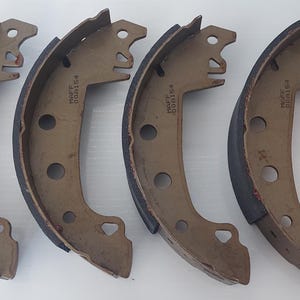 May include: Four new brake shoes, arranged in a row, with a curved, metallic brown body and a black friction material. The shoes have multiple holes and a distinctive shape. An orange box with text is partially visible in the background.