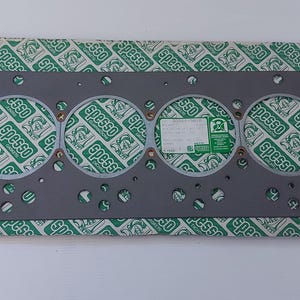 Puede incluir: Una junta de culata gris con seis aberturas circulares y numerosos agujeros m&aacute;s peque&ntilde;os. La junta est&aacute; sobre un papel estampado verde y blanco con la palabra "Spesso" repetida. Una etiqueta blanca y verde est&aacute; adjunta.