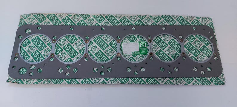 Puede incluir: Una junta de culata gris con seis aberturas circulares y numerosos agujeros m&aacute;s peque&ntilde;os. La junta est&aacute; sobre un papel estampado verde y blanco con la palabra "Spesso" repetida. Una etiqueta blanca y verde est&aacute; adjunta.