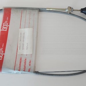 Puede incluir: Un cable plateado enrollado con un botón negro y un paquete de plástico transparente con detalles rojos. El cable es probablemente un componente mecánico, posiblemente para un vehículo. El paquete tiene texto en un idioma extranjero.