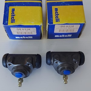 Può includere: Due cilindri freno per autoveicoli con dettagli blu e raccordi in ottone sono esposti sotto due scatole Metelli gialle e blu. Le scatole riportano la scritta "metelli" e "04-0318".