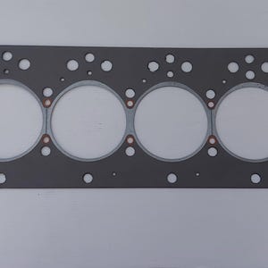 Puede incluir: Una junta de culata rectangular gris con seis grandes aberturas circulares y numerosos agujeros m&aacute;s peque&ntilde;os. Se ven anillos de color cobre alrededor de las aberturas circulares. La junta est&aacute; dise&ntilde;ada para un motor.