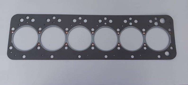 Puede incluir: Una junta de culata rectangular gris con seis grandes aberturas circulares y numerosos agujeros m&aacute;s peque&ntilde;os. Se ven anillos de color cobre alrededor de las aberturas circulares. La junta est&aacute; dise&ntilde;ada para un motor.