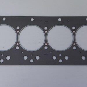 Puede incluir: Una junta de culata gris con seis grandes aberturas circulares y numerosos agujeros m&aacute;s peque&ntilde;os. La junta es rectangular con esquinas redondeadas y un interior de color claro. La imagen es una toma de cerca de la junta.