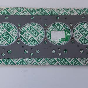 Puede incluir: Una junta de culata gris con seis grandes aberturas circulares y numerosos agujeros m&aacute;s peque&ntilde;os. La junta est&aacute; sobre un papel estampado verde y blanco con la palabra "spesso" repetida. Se adjunta una peque&ntilde;a etiqueta blanca.