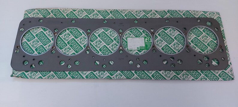 Puede incluir: Una junta de culata gris con seis grandes aberturas circulares y numerosos agujeros m&aacute;s peque&ntilde;os. La junta est&aacute; sobre un papel estampado verde y blanco con la palabra "spesso" repetida. Se adjunta una peque&ntilde;a etiqueta blanca.