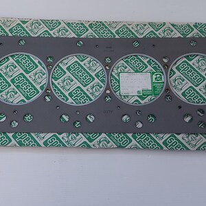 Puede incluir: Una junta de culata de metal gris con seis grandes aberturas circulares y numerosos agujeros m&aacute;s peque&ntilde;os. La junta descansa sobre un papel estampado verde y blanco con la palabra "Spesso" repetida. Una peque&ntilde;a etiqueta blanca es visible.