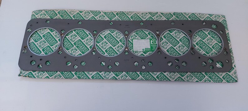 Puede incluir: Una junta de culata de metal gris con seis grandes aberturas circulares y numerosos agujeros m&aacute;s peque&ntilde;os. La junta descansa sobre un papel estampado verde y blanco con la palabra "Spesso" repetida. Una peque&ntilde;a etiqueta blanca es visible.