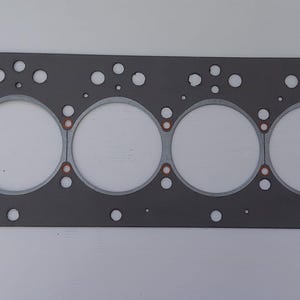 Puede incluir: Una junta de culata gris con seis grandes aberturas circulares y numerosos agujeros m&aacute;s peque&ntilde;os. La junta tiene anillos de color cobre alrededor de las aberturas grandes. La forma rectangular sugiere un componente automotriz o de motor.