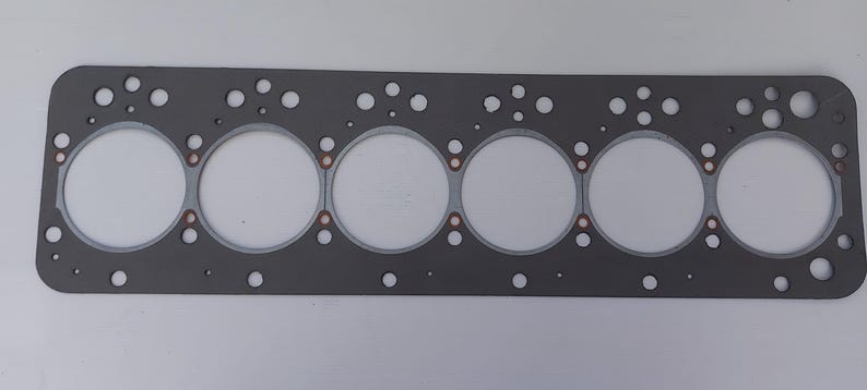 Puede incluir: Una junta de culata gris con seis grandes aberturas circulares y numerosos agujeros m&aacute;s peque&ntilde;os. La junta tiene anillos de color cobre alrededor de las aberturas grandes. La forma rectangular sugiere un componente automotriz o de motor.