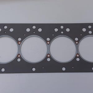 Puede incluir: Una junta de culata rectangular gris con seis grandes aberturas circulares y numerosos agujeros m&aacute;s peque&ntilde;os. La junta tiene detalles de color cobre alrededor de las aberturas circulares. Dise&ntilde;ada para reparaci&oacute;n de autom&oacute;viles o motores.