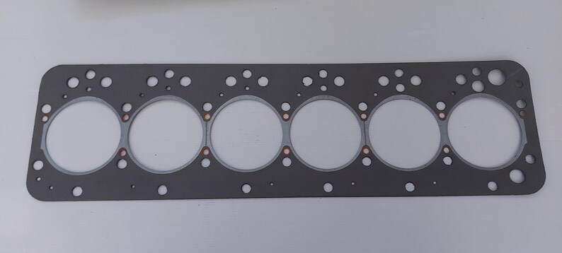 Puede incluir: Una junta de culata rectangular gris con seis grandes aberturas circulares y numerosos agujeros m&aacute;s peque&ntilde;os. La junta tiene detalles de color cobre alrededor de las aberturas circulares. Dise&ntilde;ada para reparaci&oacute;n de autom&oacute;viles o motores.