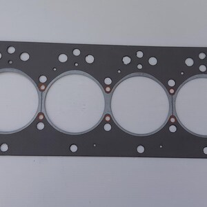 Puede incluir: Una junta de culata de motor gris con seis grandes aberturas circulares y numerosos agujeros m&aacute;s peque&ntilde;os. La junta tiene forma rectangular con esquinas redondeadas y detalles cobrizos alrededor de las aberturas circulares. Para automoci&oacute;n.