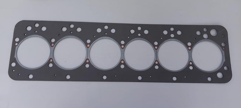 Puede incluir: Una junta de culata de motor gris con seis grandes aberturas circulares y numerosos agujeros m&aacute;s peque&ntilde;os. La junta tiene forma rectangular con esquinas redondeadas y detalles cobrizos alrededor de las aberturas circulares. Para automoci&oacute;n.