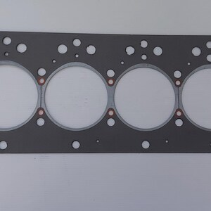 Puede incluir: Una junta de culata gris con seis grandes aberturas circulares y numerosos agujeros m&aacute;s peque&ntilde;os. La junta tiene forma rectangular con esquinas redondeadas y detalles en color cobre. Dise&ntilde;ada para reparaci&oacute;n de autom&oacute;viles o motores.