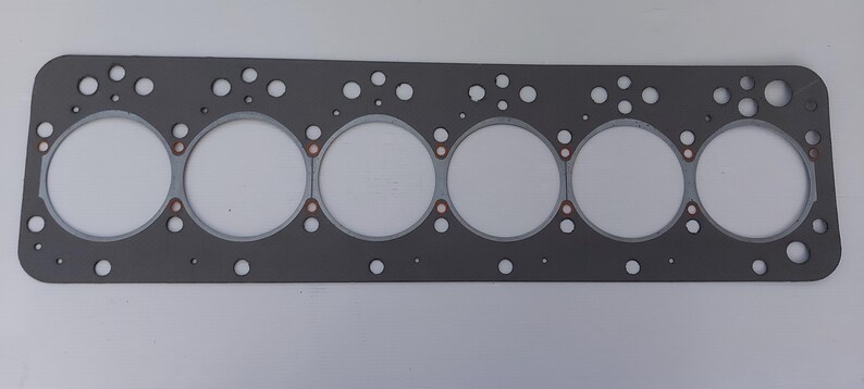 Puede incluir: Una junta de culata gris con seis grandes aberturas circulares y numerosos agujeros m&aacute;s peque&ntilde;os. La junta tiene forma rectangular con esquinas redondeadas y detalles en color cobre. Dise&ntilde;ada para reparaci&oacute;n de autom&oacute;viles o motores.