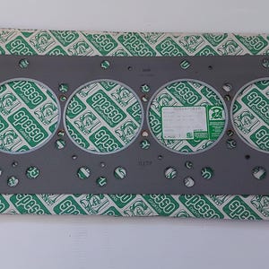 Puede incluir: Una junta de culata gris con seis grandes aberturas circulares y numerosos agujeros m&aacute;s peque&ntilde;os. La junta descansa sobre un papel estampado verde y blanco con la palabra "Spesso" impresa. Una etiqueta es visible.