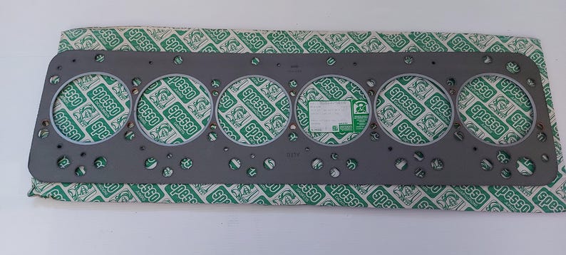 Puede incluir: Una junta de culata gris con seis grandes aberturas circulares y numerosos agujeros m&aacute;s peque&ntilde;os. La junta descansa sobre un papel estampado verde y blanco con la palabra "Spesso" impresa. Una etiqueta es visible.