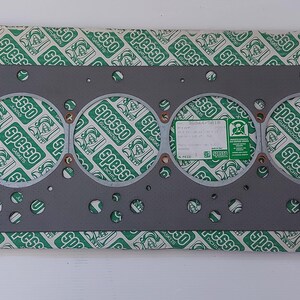 Puede incluir: Una junta de culata gris con seis grandes aberturas circulares y numerosos agujeros m&aacute;s peque&ntilde;os. La junta est&aacute; sobre un papel estampado verde y blanco con la palabra "Spesso" repetida. Una peque&ntilde;a etiqueta es visible.