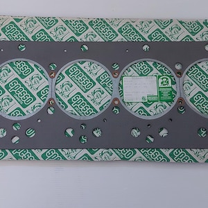 Puede incluir: Una junta de culata gris con seis grandes aberturas circulares y numerosos agujeros m&aacute;s peque&ntilde;os. La junta descansa sobre un papel estampado en verde y blanco con la palabra "Spesso" repetida. Una peque&ntilde;a etiqueta es visible.