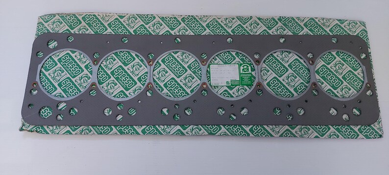 Puede incluir: Una junta de culata gris con seis grandes aberturas circulares y numerosos agujeros m&aacute;s peque&ntilde;os. La junta descansa sobre un papel estampado en verde y blanco con la palabra "Spesso" repetida. Una peque&ntilde;a etiqueta es visible.
