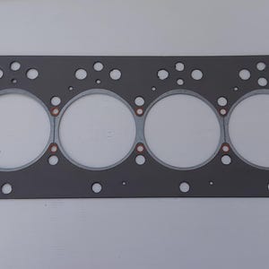 Puede incluir: Una junta de culata rectangular gris con seis grandes aberturas circulares y numerosos agujeros m&aacute;s peque&ntilde;os. Se ven anillos de color cobre alrededor de las aberturas grandes. La junta est&aacute; dise&ntilde;ada para aplicaciones automotrices o de motor.