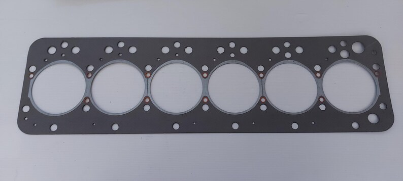 Puede incluir: Una junta de culata rectangular gris con seis grandes aberturas circulares y numerosos agujeros m&aacute;s peque&ntilde;os. Se ven anillos de color cobre alrededor de las aberturas grandes. La junta est&aacute; dise&ntilde;ada para aplicaciones automotrices o de motor.