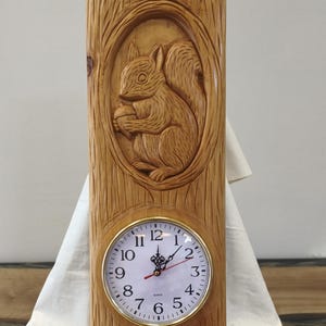 Puede incluir: Un reloj de pared rectangular de madera con una ardilla tallada a mano sosteniendo una bellota dentro de un marco ovalado. La esfera es blanca con números arábigos negros, un borde dorado y manecillas negras con un segundero de color rojo.