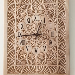 Puede incluir: Un reloj cuadrado de madera con un intrincado diseño tallado. La esfera del reloj presenta números romanos y manecillas. El marco y la esfera del reloj están tallados con un patrón repetitivo de hojas y formas geométricas. El reloj está hecho de madera clara.