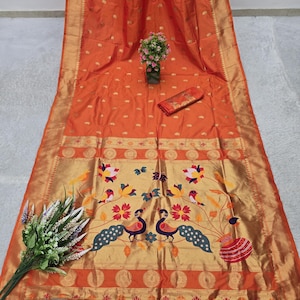 Puede incluir: Un sari de seda naranja con ribete dorado y un borde decorativo con pavos reales, pájaros y diseños florales. El sari tiene una pieza de tela a juego y está adornado con borlas naranjas.