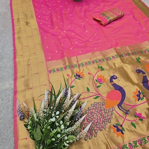 K&ouml;nnte beinhalten: Ein leuchtend rosa Sari mit goldenem Rand und Pfauenmuster. Der Sari hat ein gold-rosa Blumenmuster und ein passendes Stoffst&uuml;ck. Ein Blumenstrau&szlig; und eine kleine Topfpflanze sind ebenfalls vorhanden.