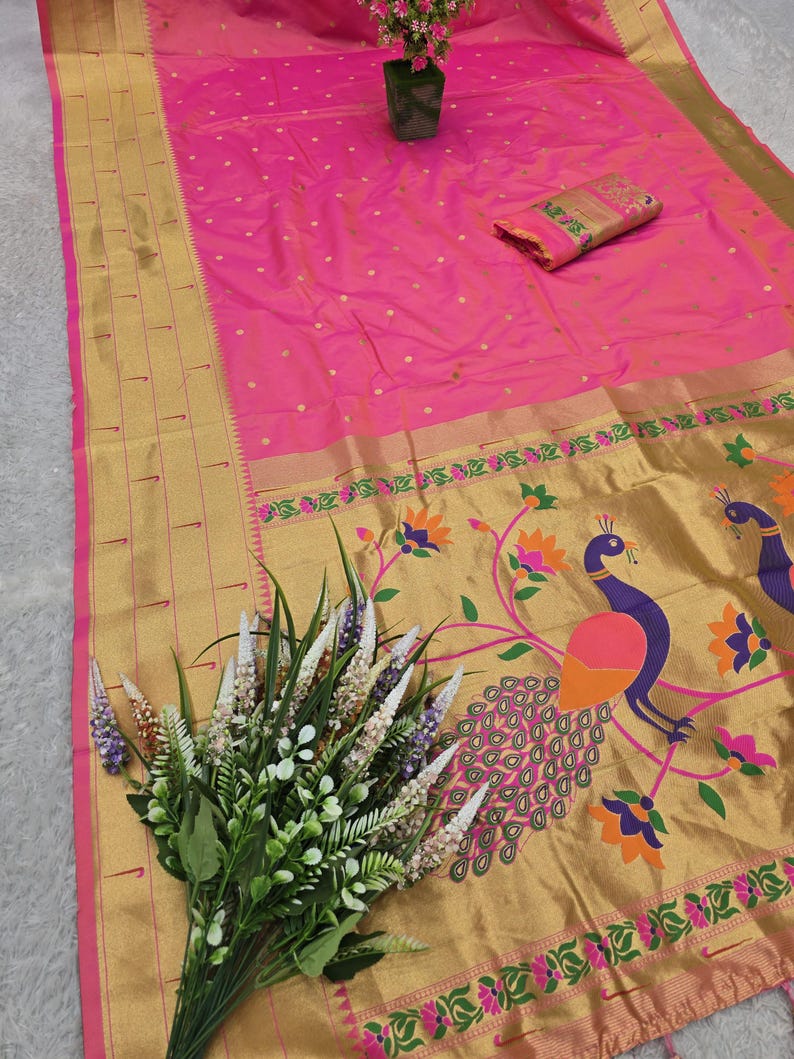 K&ouml;nnte beinhalten: Ein leuchtend rosa Sari mit goldenem Rand und Pfauenmuster. Der Sari hat ein gold-rosa Blumenmuster und ein passendes Stoffst&uuml;ck. Ein Blumenstrau&szlig; und eine kleine Topfpflanze sind ebenfalls vorhanden.