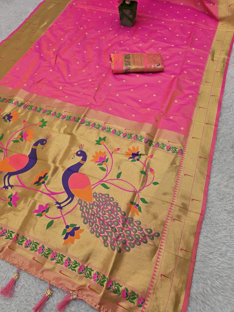 K&ouml;nnte beinhalten: Ein leuchtend rosa Sari mit goldenem Rand und Pfauenmuster. Der Sari hat einen detaillierten Rand mit Blumen- und Pfauenmotiven. Ein passendes Stoffst&uuml;ck ist enthalten. Der Sari ist gefaltet und auf einer hellen Oberfl&auml;che ausgestellt.