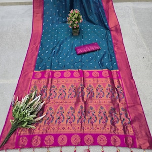 May include: A teal-colored sari with a pink border and intricate floral patterns. The sari features a detailed design along the bottom edge with tassels. A small pink clutch and a small potted plant are also visible.