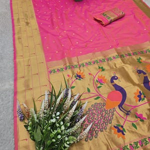 K&ouml;nnte beinhalten: Ein leuchtend pinkfarbener Sari mit goldenem Rand und Pfauenmuster. Der Sari hat ein gold-rosa Blumenmuster und ein passendes Stoffst&uuml;ck. Ein Blumenstrau&szlig; und eine kleine Topfpflanze sind ebenfalls vorhanden.
