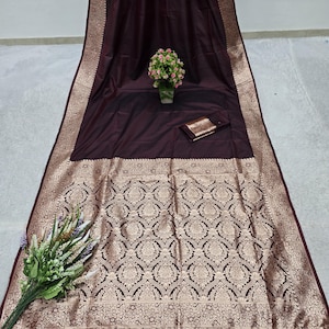 May include: A maroon saree with a detailed gold pattern on the lower section and border. The saree has a matching maroon fringe and a small matching pouch. A vase of pink flowers and a bouquet of flowers are also present.