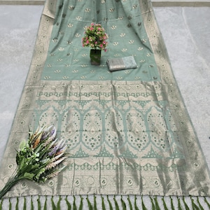 Op de afbeelding: Een lichtgroene sari met ingewikkelde goudkleurige bloemen- en paisley-patronen. De sari heeft een decoratieve rand en groene kwastjes langs de rand. Een klein boeket bloemen en een opgerold stuk stof zijn ook zichtbaar.