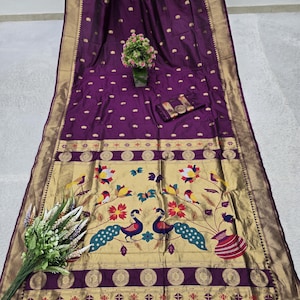 Puede incluir: Un lujoso sari de seda púrpura con detalles dorados y un borde detallado. El sari presenta un panel decorativo con motivos de pavo real y florales en colores vibrantes. Las borlas cuelgan del borde inferior, añadiendo un toque de elegancia.