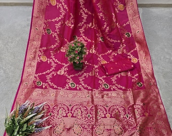 May include: A magenta-colored saree with intricate gold floral patterns and a decorative border. The saree features a matching blouse piece and is adorned with tassels along the bottom edge. A small plant and a bouquet of flowers are also present.
