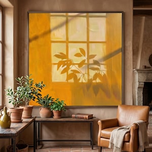 May include: A large abstract painting in yellow and orange tones shows a window with leaf shadows. The artwork hangs above a dark metal table and a leather armchair. Potted plants and a book are also visible in the room.