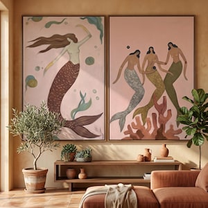 May include: Two framed paintings of mermaids. One shows a mermaid with brown hair and a brown tail, dancing against a pink backdrop. The second depicts three mermaids with silver, gold, and green tails, holding hands. Coral and seaweed are also depicted.