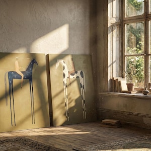 May include: Two canvas art prints leaning against a wall. One features a dark blue horse with a rider, the other a white horse with black spots. Both have elongated legs and are set against a muted background. The room has a rustic feel with a window and sunlight.