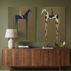 May include: Two framed art prints featuring stylized horses with riders. One horse is dark blue with star-like patterns, the other is white with black spots. The artwork is displayed above a wooden cabinet with a lamp and decorative objects.