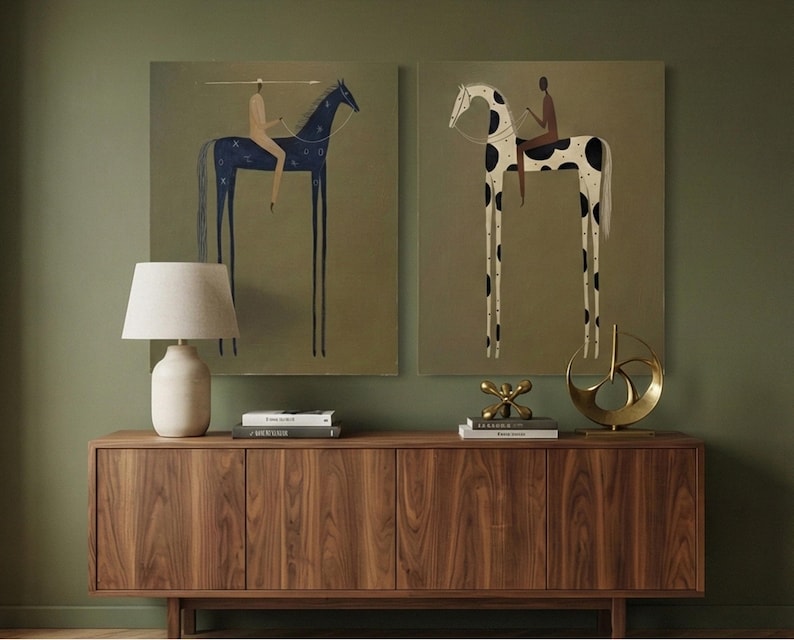 May include: Two framed art prints featuring stylized horses with riders. One horse is dark blue with star-like patterns, the other is white with black spots. The artwork is displayed above a wooden cabinet with a lamp and decorative objects.
