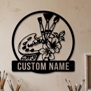 May include: Black metal wall art featuring a painter's palette, paintbrushes, and flowers within a circular frame. The text "CUSTOM NAME" is displayed on a rectangular plaque below the design. The art is displayed on a beige wall.