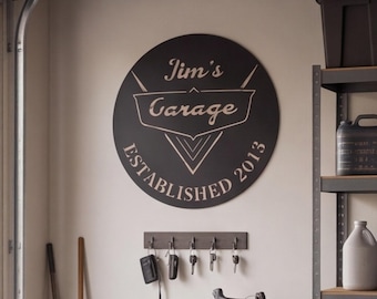 以下が含まれることがあります： Jim's GarageおよびESTABLISHED 2013という白い文字と中央のグラフィックエンブレムが特徴の円形の黒い壁掛け看板。看板の下には、鍵や黒いキーフォブを掛けた5つのフックが付いた長方形の壁掛けキーラックがあります。