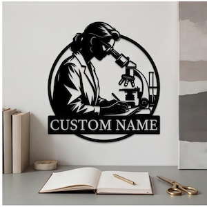 Può includere: Un'opera d'arte da parete in metallo nero che raffigura la silhouette di uno scienziato che guarda attraverso un microscopio. Il design circolare include un banner rettangolare nella parte inferiore con il testo CUSTOM NAME in lettere bianche.