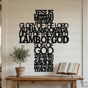 Könnte beinhalten: Ein schwarzes Metallkreuz mit religiösen Texten, darunter "JESUS", "LAMB OF GOD" und "SON OF GOD". Das Kreuz ist an einer weißen Wand über einem Holztisch angebracht. Ein offenes Buch und eine Topfpflanze stehen auf dem Tisch.